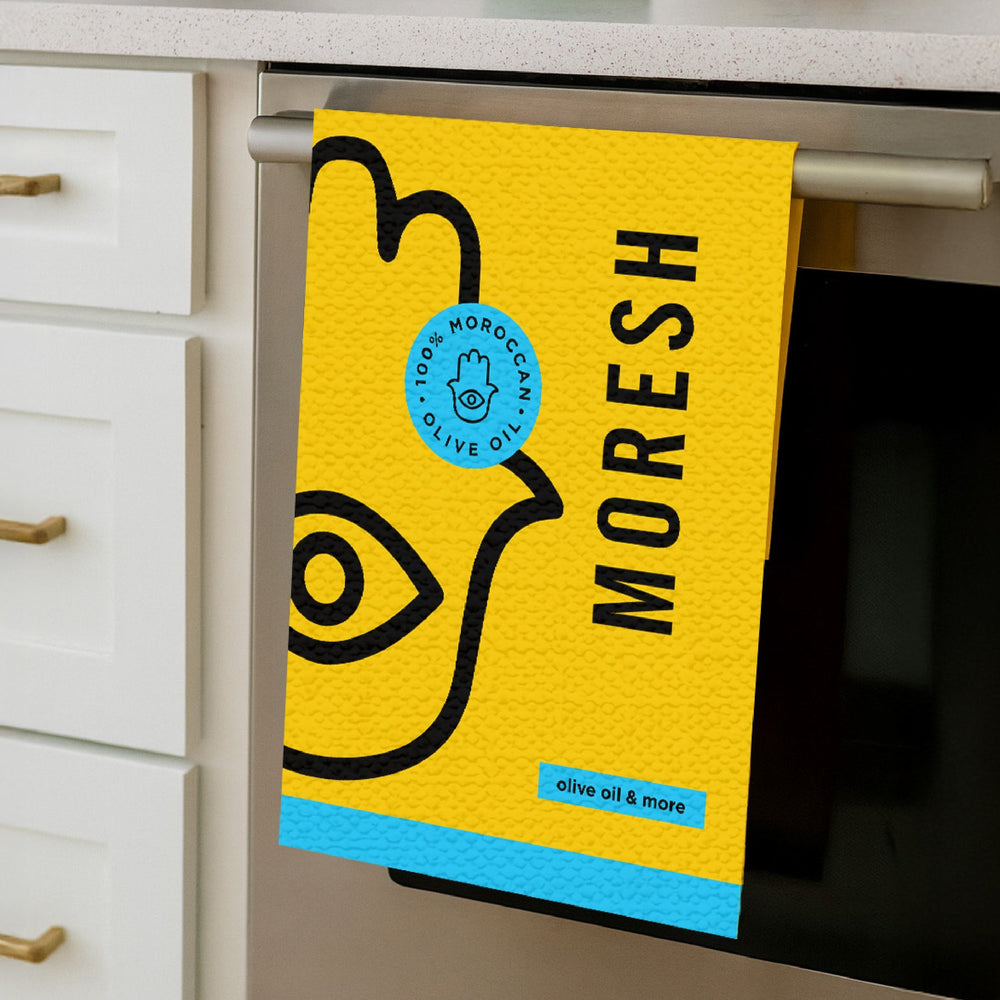 Moresh branded tea towel in yellow, hanging neatly on an oven handle, showcasing its soft cotton texture and bold logo design.