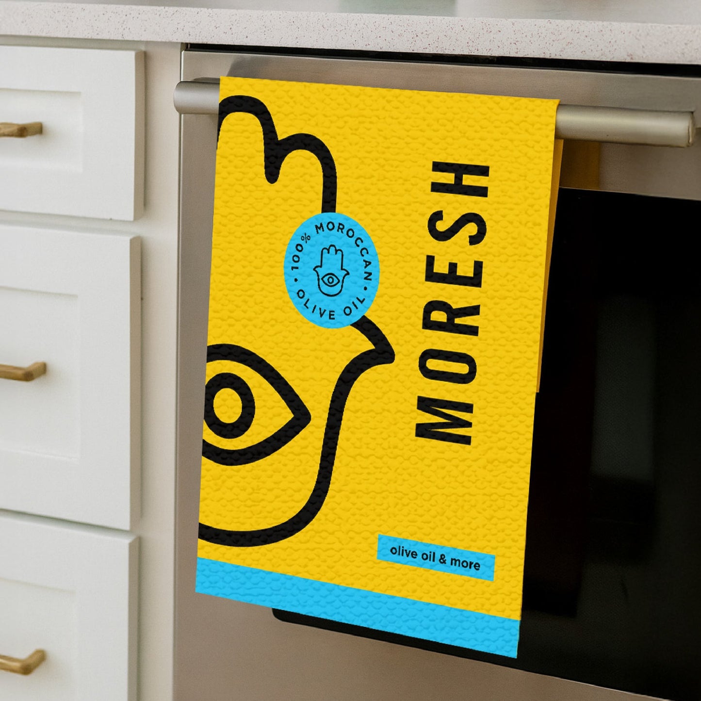 Moresh branded tea towel in yellow, hanging neatly on an oven handle, showcasing its soft cotton texture and bold logo design.